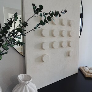 Peut inclure: Une &oelig;uvre d'art murale textur&eacute;e blanc cass&eacute; avec une s&eacute;rie d'accents circulaires en relief. Une branche d'eucalyptus vert et un vase d&eacute;coratif sont au premier plan. L'&oelig;uvre est expos&eacute;e sur une surface noire.