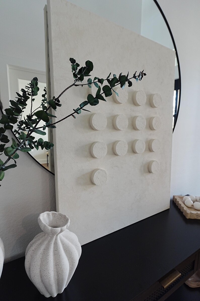 Peut inclure: Une &oelig;uvre d'art murale textur&eacute;e blanc cass&eacute; avec une s&eacute;rie d'accents circulaires en relief. Une branche d'eucalyptus vert et un vase d&eacute;coratif sont au premier plan. L'&oelig;uvre est expos&eacute;e sur une surface noire.