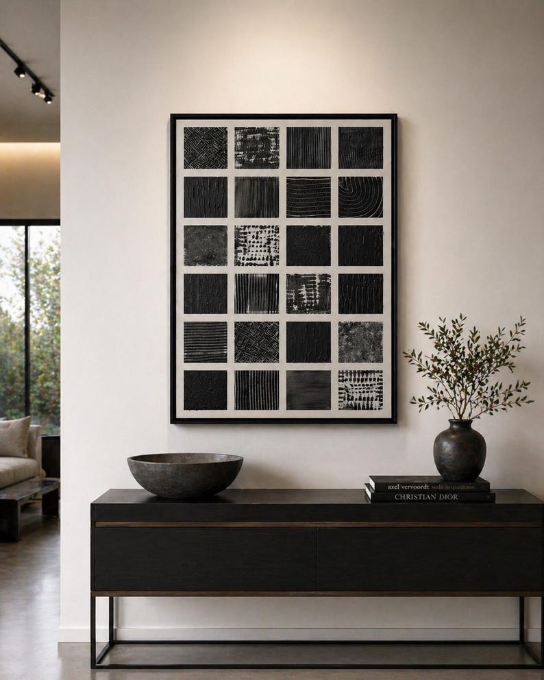 May include: A framed wall art piece featuring a grid of twenty-four black squares with varied textures on a white background. The artwork is displayed above a black console table holding a dark bowl, a vase with greenery, and two stacked books.