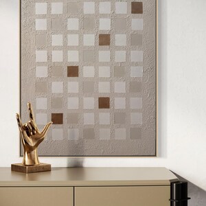 K&ouml;nnte beinhalten: Abstrakte Wandkunst mit einem Raster aus quadratischen Feldern in Beige- und Braunt&ouml;nen. Auf einem beigefarbenen Tisch steht eine goldfarbene Skulptur einer Hand, die das 'Ich liebe dich'-Zeichen macht. Das Kunstwerk ist in einem hellbraunen Rahmen gerahmt.