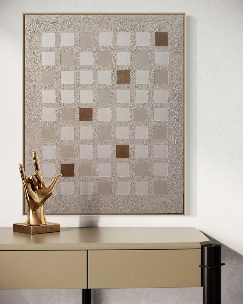 K&ouml;nnte beinhalten: Abstrakte Wandkunst mit einem Raster aus quadratischen Feldern in Beige- und Braunt&ouml;nen. Auf einem beigefarbenen Tisch steht eine goldfarbene Skulptur einer Hand, die das 'Ich liebe dich'-Zeichen macht. Das Kunstwerk ist in einem hellbraunen Rahmen gerahmt.
