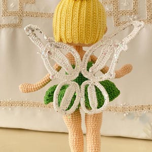 May include: A crocheted fairy doll with yellow hair, a green dress, and white wings with glitter. The doll is standing on a white surface.