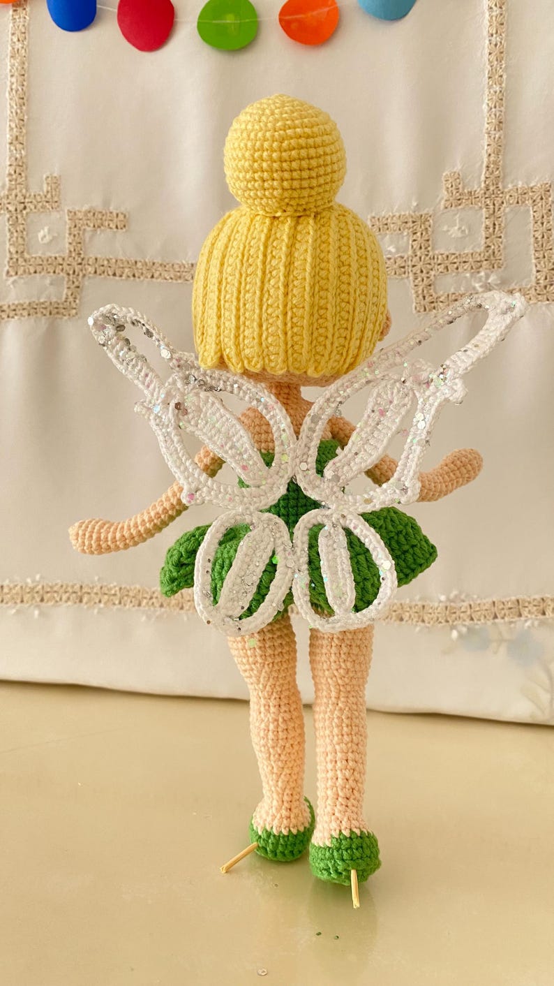 May include: A crocheted fairy doll with yellow hair, a green dress, and white wings with glitter. The doll is standing on a white surface.