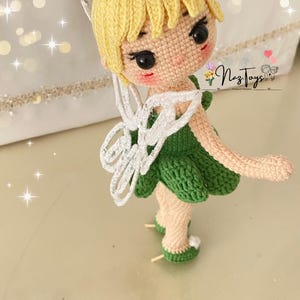 May include: A crocheted fairy doll with blonde hair, a green dress, white wings, and green shoes. The doll is standing on a white surface. The text "Naz Toys" is visible in the background.