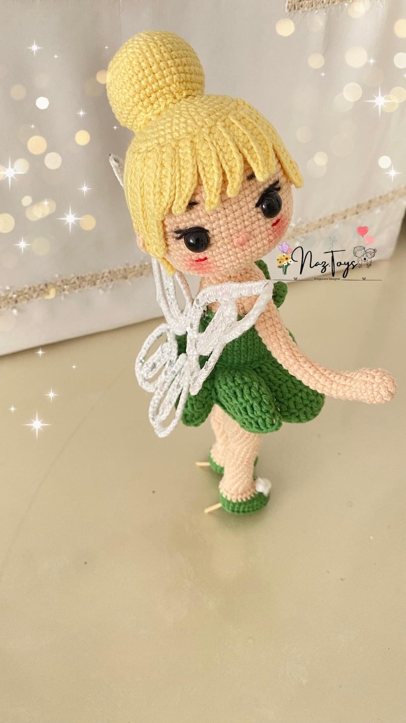May include: A crocheted fairy doll with blonde hair, a green dress, white wings, and green shoes. The doll is standing on a white surface. The text "Naz Toys" is visible in the background.