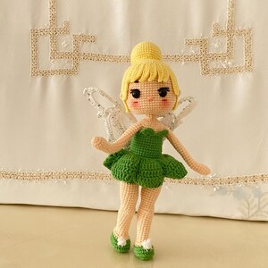 May include: A crocheted fairy doll with yellow hair, a green dress, and white wings. The doll is standing on a white surface with a colorful garland in the background.