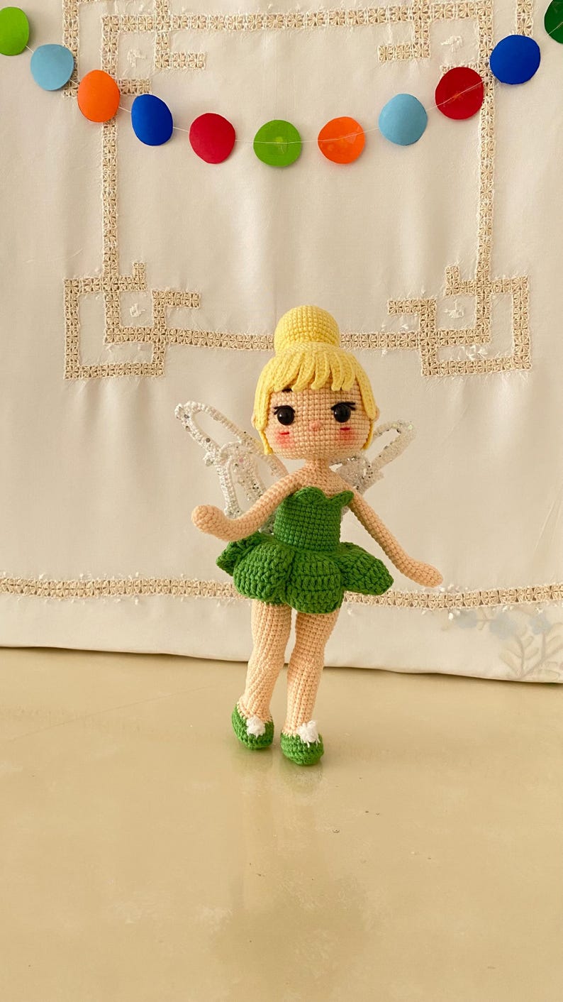 May include: A crocheted fairy doll with yellow hair, a green dress, and white wings. The doll is standing on a white surface with a colorful garland in the background.