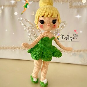 May include: A crocheted Tinkerbell doll with a green dress, yellow hair, and sparkly wings. The doll is standing on a white surface with a blurred background.