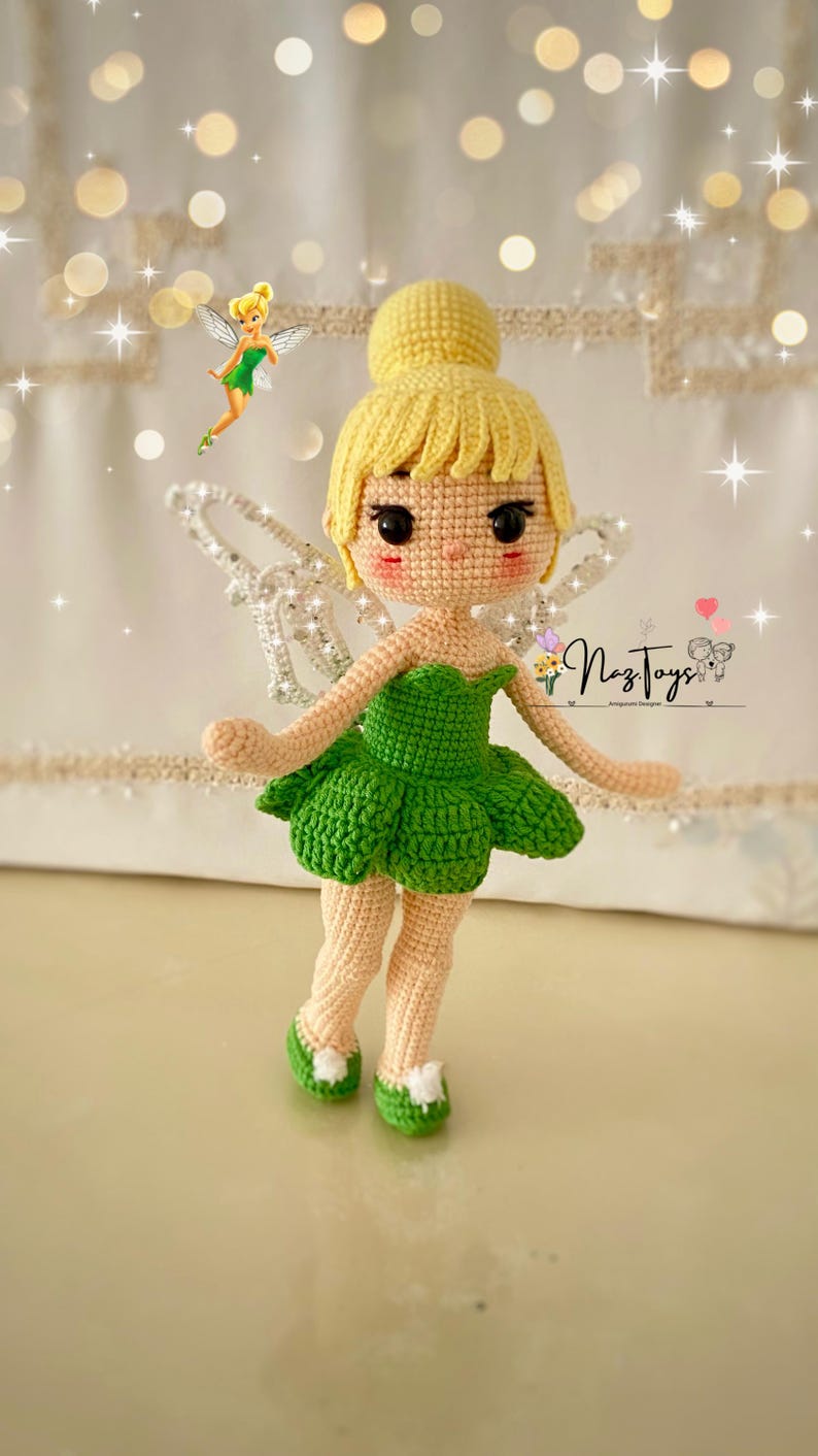 May include: A crocheted Tinkerbell doll with a green dress, yellow hair, and sparkly wings. The doll is standing on a white surface with a blurred background.