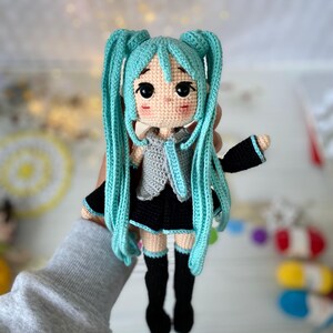 May include: A handcrafted crochet doll featuring long, bright turquoise hair styled in twin tails. The doll has a light beige complexion, large black eyes, and rosy cheeks. It wears a grey sleeveless top, a black skirt, and black knee-high socks.