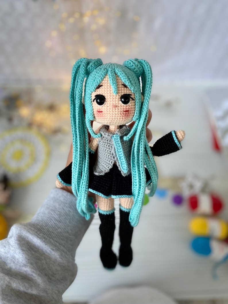 May include: A handcrafted crochet doll featuring long, bright turquoise hair styled in twin tails. The doll has a light beige complexion, large black eyes, and rosy cheeks. It wears a grey sleeveless top, a black skirt, and black knee-high socks.