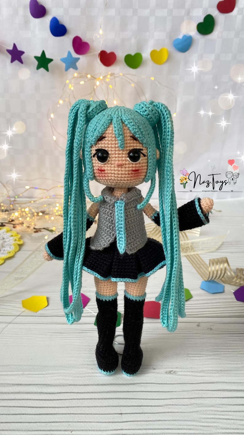 May include: A crocheted doll with long turquoise hair in twin tails, wearing a gray sleeveless top with a turquoise tie, a black pleated skirt, black arm warmers with turquoise trim, and black knee-high boots. The doll has large black eyes.