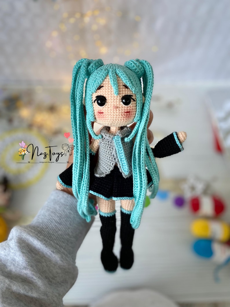 May include: A crocheted doll featuring long turquoise hair in twin tails, large black eyes, and rosy cheeks. The doll wears a grey collared top, a black skirt with turquoise trim, and black knee-high socks. A logo reading Naz Toys is visible.