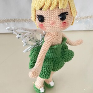 May include: A crocheted fairy doll with yellow hair, green dress, and white wings. The doll has a sweet expression and is wearing green shoes.