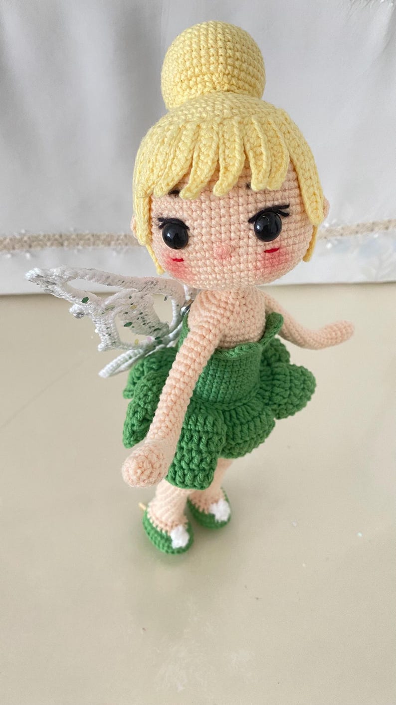 May include: A crocheted fairy doll with yellow hair, green dress, and white wings. The doll has a sweet expression and is wearing green shoes.
