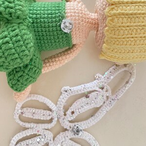 May include: A crocheted doll with a green dress, pink arms and legs, and yellow hair. The doll is missing its wings, which are shown separately in white with silver glitter.