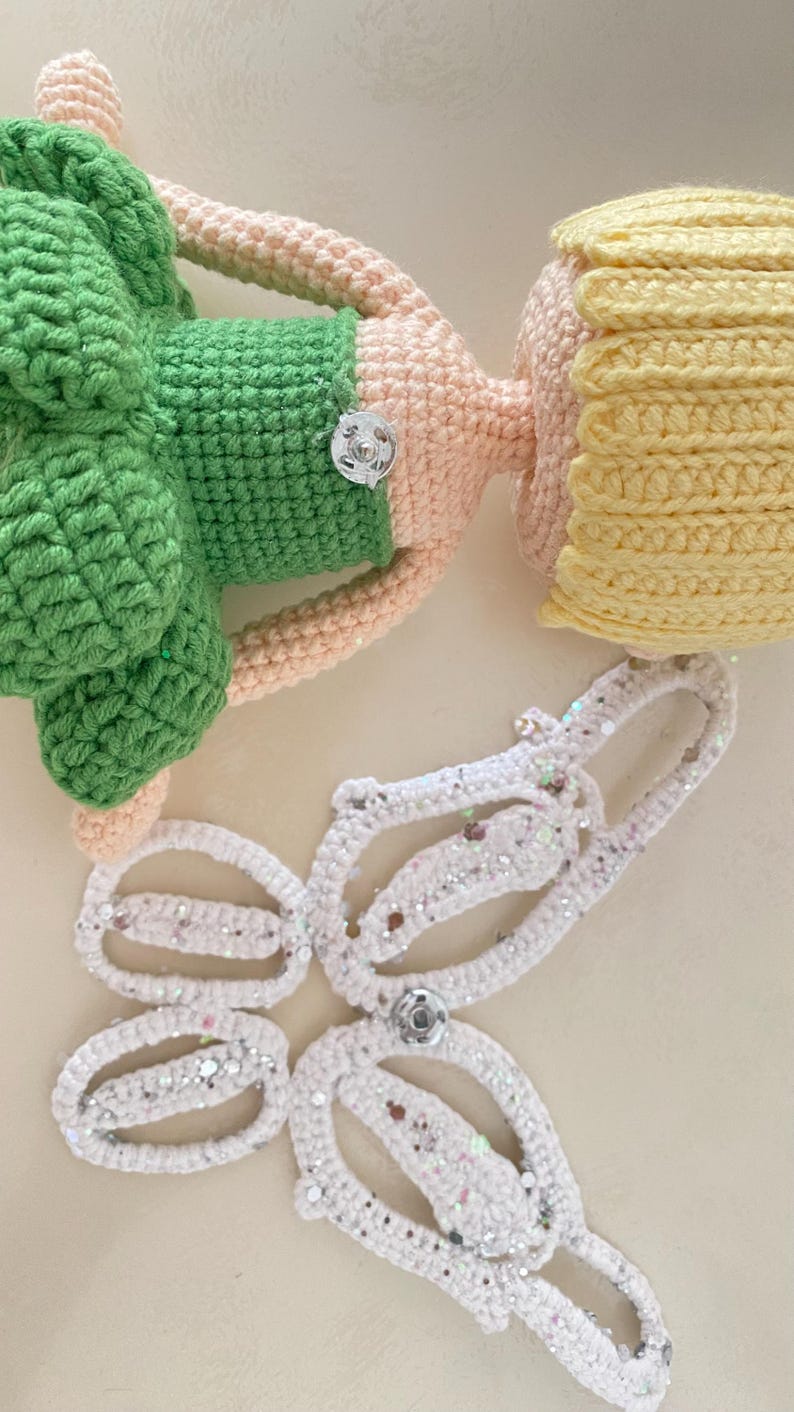 May include: A crocheted doll with a green dress, pink arms and legs, and yellow hair. The doll is missing its wings, which are shown separately in white with silver glitter.