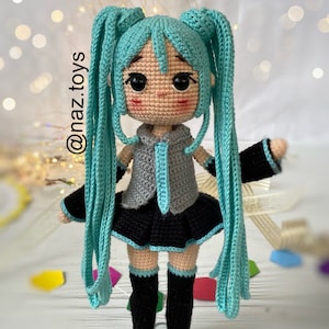 May include: A crocheted doll with long turquoise twin tails, wearing a grey sleeveless top with a matching tie, a black pleated skirt, and black knee-high boots. The doll has large black eyes and pink cheeks. The text @naz.toys is visible.