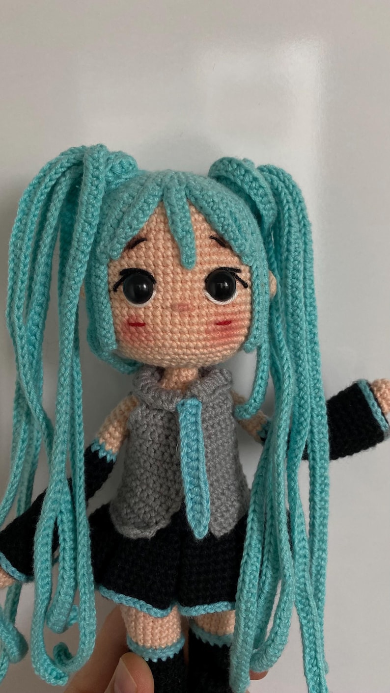 May include: A crocheted doll featuring long, bright turquoise yarn hair styled in twin tails. The doll has large black safety eyes, a small nose, and pink blush on the cheeks. It wears a gray sleeveless top with a tie, black arm warmers, and a black skirt.