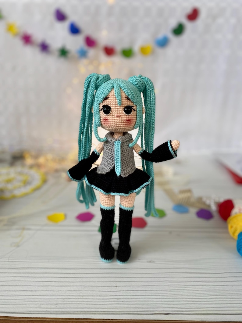 May include: A crocheted doll featuring long turquoise hair in twin tails, wearing a gray sleeveless top with a turquoise tie, a black skirt, black arm warmers, and black knee-high socks with turquoise trim. The doll has large black eyes.