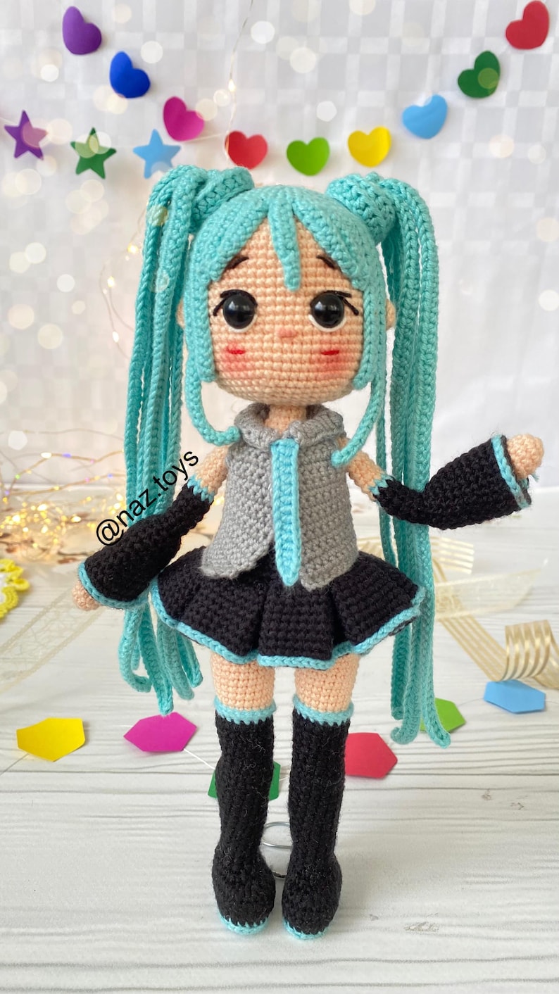 May include: A crocheted doll with long turquoise hair in twin tails, wearing a gray collared top with a turquoise tie, a black pleated skirt, black arm warmers with turquoise trim, and black knee-high boots. The doll has large black eyes and pink cheeks.