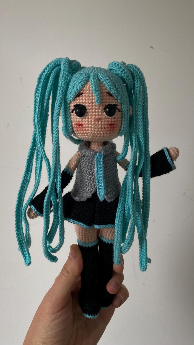 May include: A crocheted doll with long, bright turquoise yarn hair styled in twin tails. The doll has a tan complexion, large black eyes, and pink cheeks. It wears a gray sleeveless top with a turquoise tie, a black skirt, and black boots.