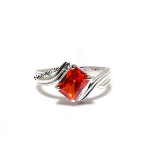 May include: A silver ring with a large, radiant, orange-red gemstone. The gemstone is square-cut and set in a modern, flowing silver band. The ring is displayed on a white background. The text "Heaven Jewelry" is visible on the ring.
