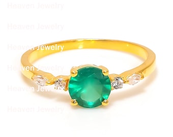 May include: A gold ring featuring a round, vibrant green gemstone as the centerpiece, flanked by smaller, clear accent stones. The ring has a delicate band and is set against a white background, showcasing the jewelry's design.