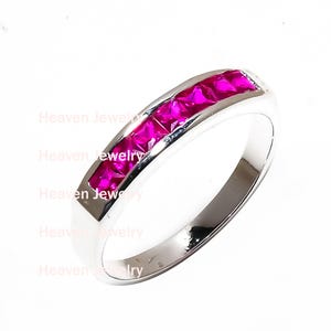 May include: A silver ring with a channel setting of vibrant pink gemstones. The ring features a polished, smooth band and a row of square-cut stones, creating a continuous line of color. The gemstones are a bright, fuchsia hue.