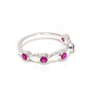 May include: A silver ring with a series of bright magenta gemstones. The ring's band has a flowing design, with each gemstone set in a bezel. The ring is shown against a white background, highlighting its elegant simplicity. A piece of jewellery.