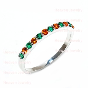 May include: A silver ring featuring a line of alternating green and orange gemstones. The ring has a shiny finish, and the gemstones are equally spaced. The words "Heaven Jewelry" are visible in the background.