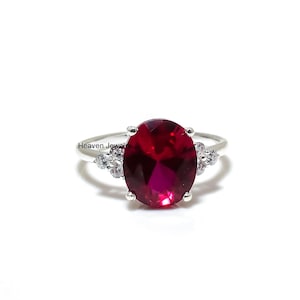 May include: A silver ring with a large, oval-cut, deep red gemstone. The ring has smaller, clear stones on either side of the main stone. The words "Heaven Jewel" are printed on the ring.