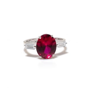 May include: A silver ring with a large, oval-cut, deep red gemstone as the focal point. Two rectangular, clear gemstones are set either side of the main stone. The ring is on a white background, with the text "Heaven Jewelry".
