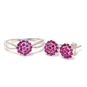 May include: A silver ring and matching stud earrings set. The ring has a split band design with a cluster of small, round, magenta gemstones. The earrings also have a cluster of magenta gemstones.