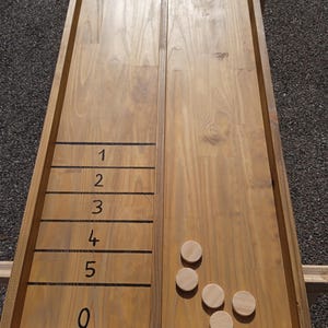 May include: A wooden shuffleboard game with a light brown finish. The board has black lines marking scoring zones from 0 to 5. Four wooden pucks are visible on the board. The game is set up on a wooden frame.