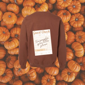 guest check with the words 'two pumpkin spice lattes please' written in dark brown handwriting font shown on the back of a brown colored crewneck