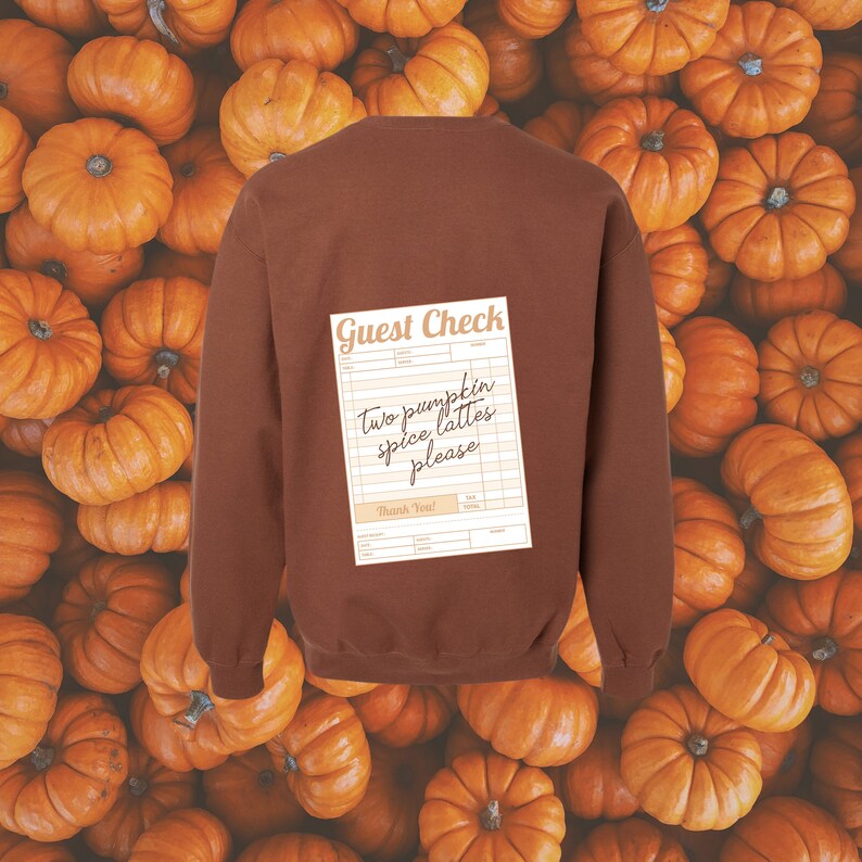 guest check with the words 'two pumpkin spice lattes please' written in dark brown handwriting font shown on the back of a brown colored crewneck