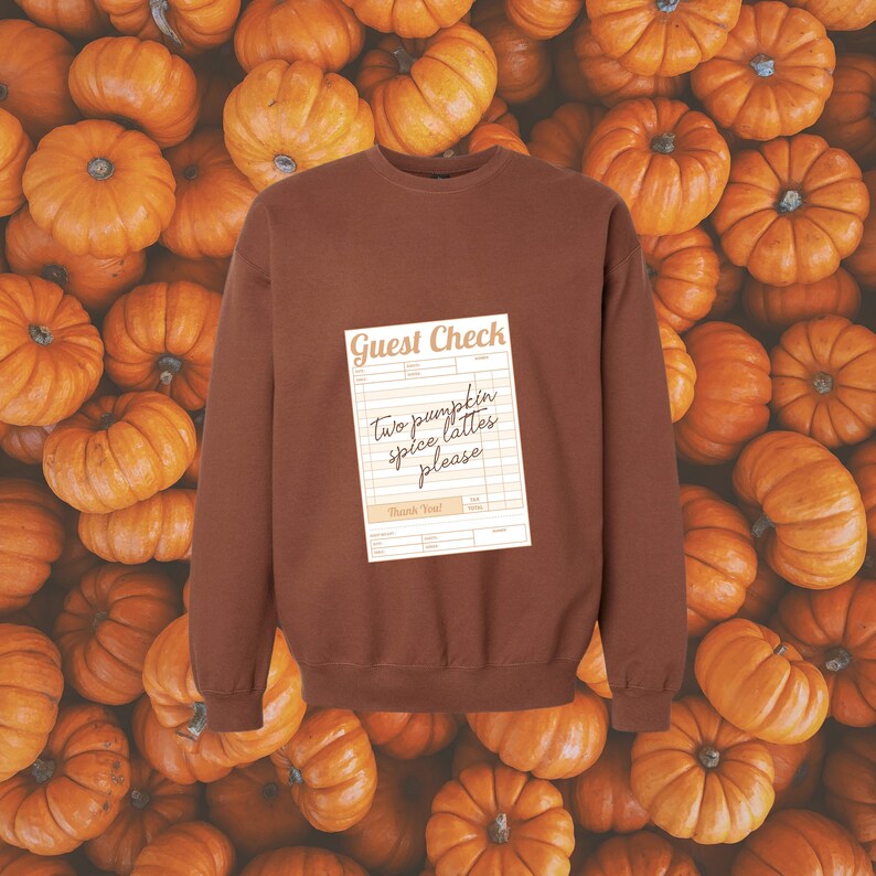 guest check with the words 'two pumpkin spice lattes please' written in dark brown handwriting font shown on the front of a brown colored crewneck