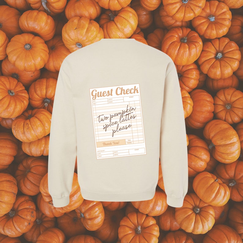 guest check with the words 'two pumpkin spice lattes please' written in dark brown handwriting font shown on the back of a cream colored crewneck