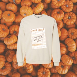 guest check with the words 'two pumpkin spice lattes please' written in dark brown handwriting font shown on the front of a cream colored crewneck