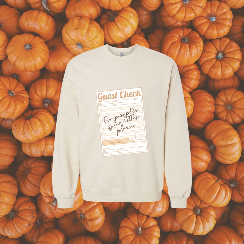 guest check with the words 'two pumpkin spice lattes please' written in dark brown handwriting font shown on the front of a cream colored crewneck