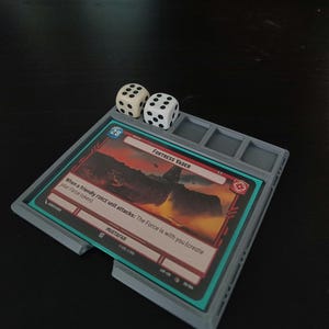 Könnte beinhalten: Ein grauer 3D-gedruckter Kartenhalter mit zwei weißen Würfeln und einer Karte mit der Aufschrift "FORTRESS VADER". Die Karte hat ein Landschaftsbild und den Text "When a friendly FORCE unit attacks: The Force is with you (create your Force token)".