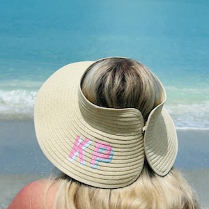 May include: A beige straw visor featuring a wide, curved brim and an open top design. The front of the visor is embroidered with the letters KP in a block font, with pink lettering outlined in light blue thread, creating a textured appearance.