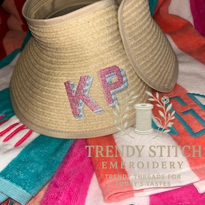 May include: A beige straw visor with embroidered pink and blue initials "KP" rests on a colorful beach towel. The towel features stripes of pink, orange, and teal. The image also includes the text "TRENDY STITCH EMBROIDERY".
