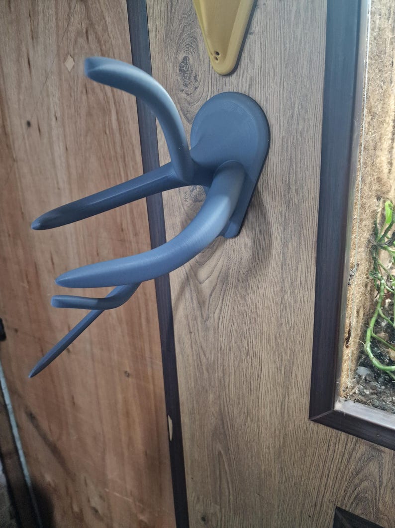 Puede incluir: Un perchero de pared de pl&aacute;stico gris en forma de cuerno de ciervo con tres ganchos. El perchero est&aacute; montado en una superficie de imitaci&oacute;n de madera.