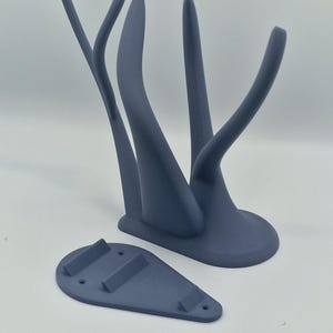 Puede incluir: Un soporte de pared de color gris impreso en 3D con un dise&ntilde;o abstracto &uacute;nico. El soporte tiene dos brazos curvos y una base que se fija a la pared.