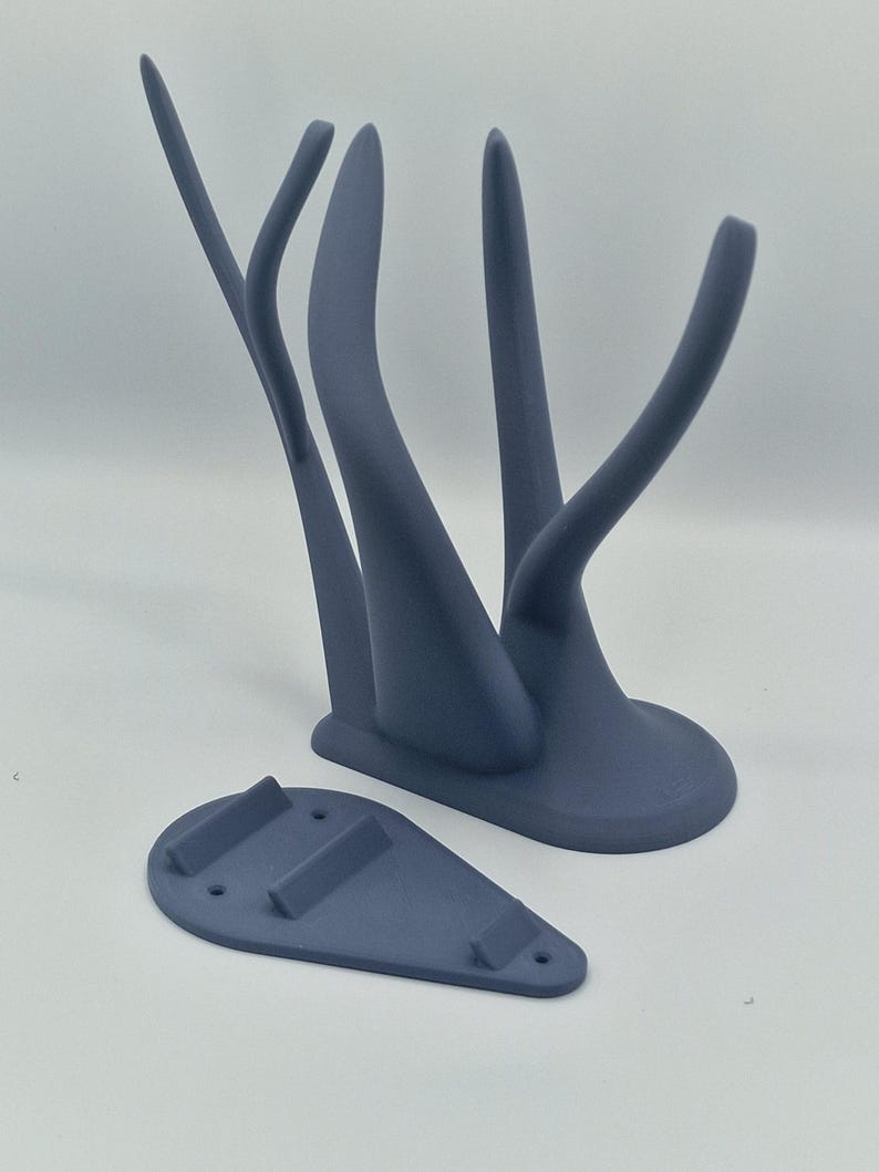 Puede incluir: Un soporte de pared de color gris impreso en 3D con un dise&ntilde;o abstracto &uacute;nico. El soporte tiene dos brazos curvos y una base que se fija a la pared.