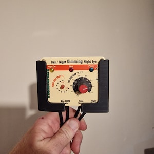 May include: A white and black thermostat with a red knob and the text "Day/Night Dimming Night Eye". The thermostat has a dial with the text "Night Time Drop °C" and "Day Time Temp °C/°F".