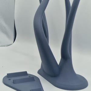 Puede incluir: Un soporte de pared de color gris impreso en 3D con dos brazos curvos y una base con tres ranuras. El soporte est&aacute; dise&ntilde;ado para sujetar art&iacute;culos como auriculares o joyas.