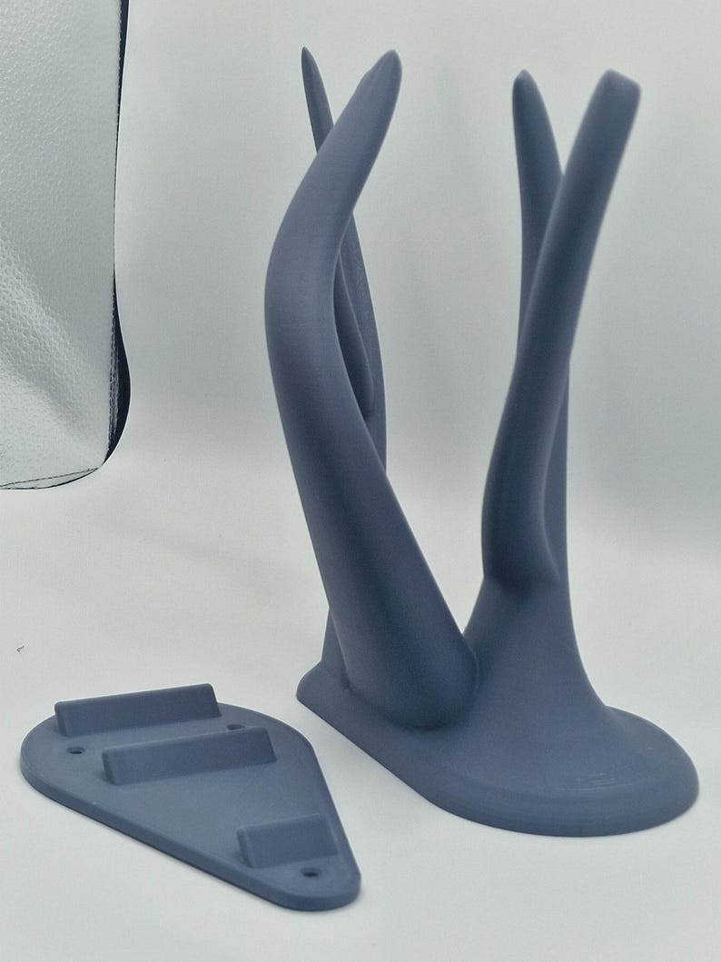 Puede incluir: Un soporte de pared de color gris impreso en 3D con dos brazos curvos y una base con tres ranuras. El soporte est&aacute; dise&ntilde;ado para sujetar art&iacute;culos como auriculares o joyas.